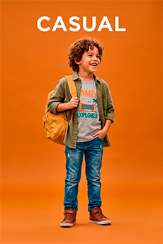 Um menino jovem com cabelo curto está sorrindo, vestindo uma camisa de flanela xadrez colorida sobre uma camiseta branca, calça jeans azul e tênis brancos. Ele está com as mãos nos bolsos da calça e posando contra um fundo amarelo.